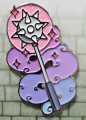 Soft enamel pin in the shape of a cleric's spiked ball mace with a handle. The spiked ball is surrounded by pink glitter to signify magic, as well as purple and blue clouds forming the background behind the handle.