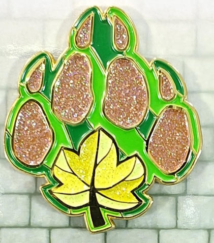 Soft enamel Druid pin in the shape of a leaf in 2 shades of green, with a brown glittery bear's paw print over the leaf. There is also a yellow leaf with some glitter st the base of the bear claw..