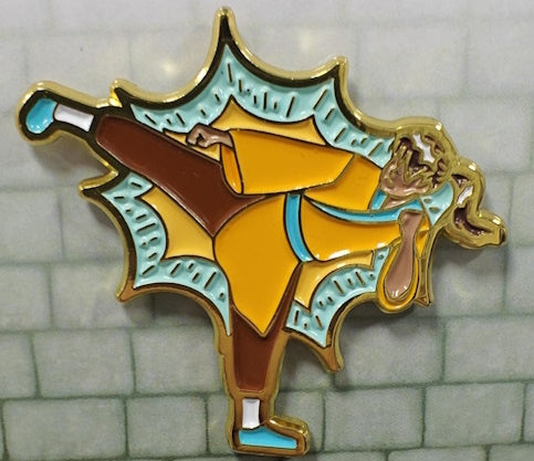 Soft enamel pin of a Monk wearing a saffron robe with teal sash and brown pants, assuming a kicking pose. It has a light teal background with gold accents depicting the force of the kick.
