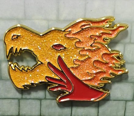 Soft enamel pin depicting a Sorcerer's hand in red with an orange and gold glittery dragon's head in the background.